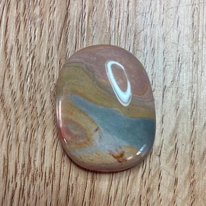 May include: A polished, oval-shaped stone with a smooth, glossy surface. The stone features layers of colour, including pink, tan, blue, and brown. The stone is approximately 5 cm long and is set against a wooden background.