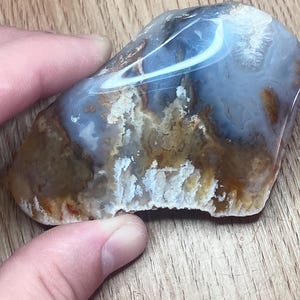 Puede incluir: Una piedra pulida de forma irregular con una mezcla de colores. La piedra presenta remolinos de azul, blanco, marrón y beige. La piedra está siendo sostenida por una mano.