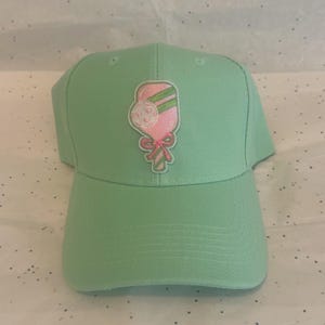 May include: A light green baseball cap with a curved brim. The cap features an embroidered design of a pink and green lollipop with a pink bow. The cap is made of a soft fabric and has a classic six-panel design.