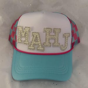 May include: A trucker hat with a white front panel and a light blue brim. The hat features the letters "MAHJ" in a pearl-like design. The sides of the hat are a pink and blue checkered pattern, with a pink rope detail.
