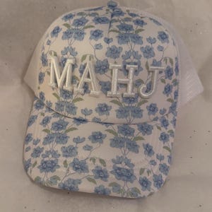 May include: A white trucker hat with a floral pattern of blue flowers and green leaves. The letters "MAHJ" are embroidered on the front in a silver color. The hat has a mesh back.