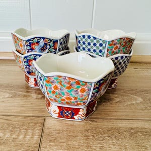 May include: A set of six decorative ceramic bowls with a scalloped rim. The bowls feature colorful floral and bird patterns in red, blue, orange, and green. The bowls are stacked on a wooden surface.