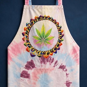 May include: A tie-dye apron featuring a pastel pink and blue swirl pattern with a central graphic. The design includes a circular ring of colorful mushrooms surrounding a detailed green cannabis leaf. The apron has two shoulder straps and a front pocket.