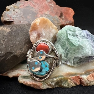 May include: A silver ring with a distinctive design, incorporating a red stone and a turquoise stone. The ring is displayed against a backdrop of various colourful rocks, including a large piece of red-orange stone.