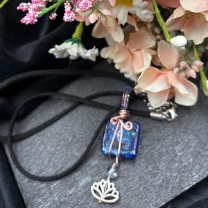 May include: A handcrafted pendant featuring a square, blue glass stone with a swirling pattern, wrapped in copper wire. A white lotus flower charm hangs below, suspended by a beaded chain. The pendant hangs from a black cord necklace.