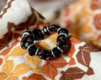 May include: A black beaded bracelet with silver-toned metal accents. The beads have a faceted design and a speckled, copper-colored pattern. The bracelet rests on a fall-themed fabric with orange, brown, and cream colors.