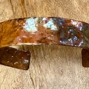 Puede incluir: Una pulsera de puño de cobre hecha a mano con un acabado texturizado e iridiscente. El diseño abierto presenta una rica mezcla de tonos marrón, bronce y verde azulado. Esta pieza de joyería única está diseñada para ser usada en la muñeca.