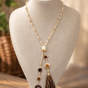 May include: A long lariat-style necklace featuring a strand of small cream and tan beads. The necklace splits into two dangling sections adorned with smooth, flat, geometric-shaped beads in shades of cream and brown, ending in a brown faux-suede tassel.