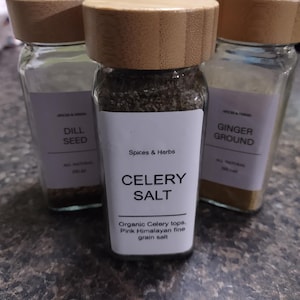 May include: Four glass spice jars with wooden lids. The jars contain dill seed, celery salt, and ginger ground. The celery salt jar label reads "Organic Celery tops. Pink Himalayan fine grain salt."