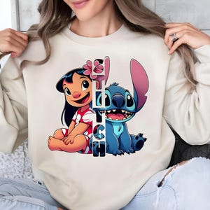 Peut inclure: Sweat-shirt crème avec un graphisme coloré de Lilo et Stitch, avec le mot "Stitch" verticalement au centre. Lilo porte une robe rouge et Stitch est bleu. Le sweat-shirt est de style décontracté.