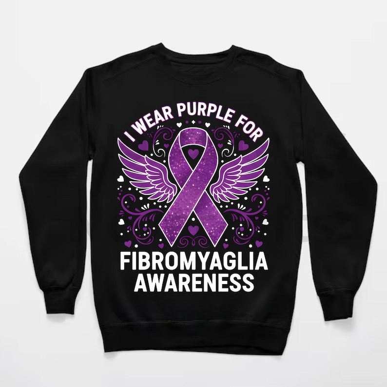 Puede incluir: Sudadera negra con un dise&ntilde;o de lazo morado, alas y el texto "I wear purple for Fibromyalgia Awareness". El dise&ntilde;o incluye corazones y adornos. Sudadera de cuello redondo.