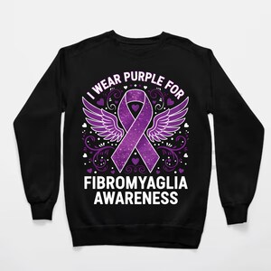 Puede incluir: Sudadera negra con un dise&ntilde;o de lazo morado, alas y el texto "I wear purple for Fibromyalgia Awareness". El dise&ntilde;o incluye corazones y adornos. Sudadera de cuello redondo.