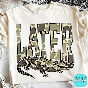 Peut inclure: T-shirt crème avec le mot "LATER" en motif camouflage au-dessus d'une illustration d'alligator. L'alligator est dans des tons de marron, vert et noir.