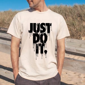 May include: A beige t-shirt with the phrase "JUST DO IT" in large, black, dripping font. The shirt is short-sleeved and appears to be made of cotton. The person is standing outdoors on a sunny day.