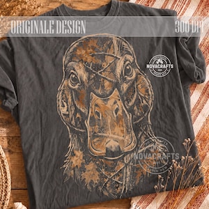 Peut inclure: Un t-shirt gris anthracite avec un grand graphique détaillé d'une tête de canard recouvert d'un motif camouflage marron de feuilles et de branches. Le texte "ORIGINALE DESIGN" figure en haut, avec des logos "NOVACRAFTS".