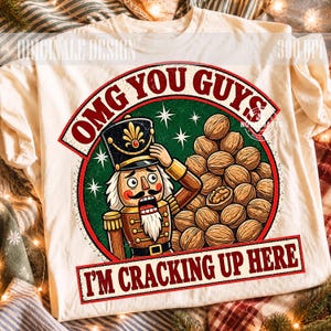 May include: A cream-colored t-shirt featuring a graphic of a nutcracker next to a pile of walnuts. The text reads OMG YOU GUYS I'M CRACKING UP HERE. The design is set within a green circle with white stars and a red border on the shirt front.