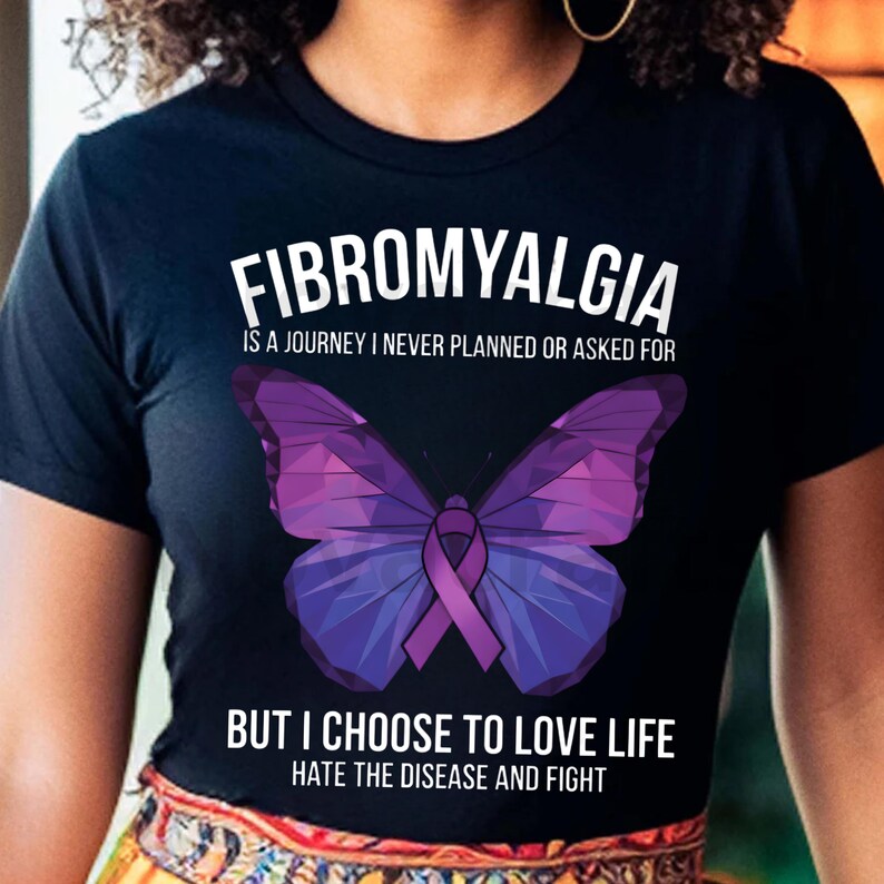 Puede incluir: Camiseta negra con la palabra "FIBROMYALGIA" sobre un dise&ntilde;o de mariposa morada. El texto debajo de la mariposa dice: "PERO ELIJO AMAR LA VIDA, ODIAR LA ENFERMEDAD Y LUCHAR".