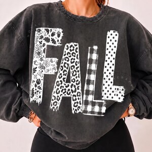 May include: A charcoal grey crewneck sweatshirt featuring the word FALL printed in large white letters. Each letter has a distinct pattern: the F has floral and acorn motifs, the A has a leopard print, the L has a buffalo check, and the L has polka dots.