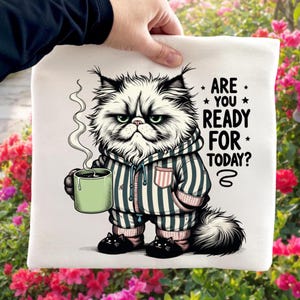 Puede incluir: Un bolso tote blanco con un gato de dibujos animados gruñón con pijama a rayas sosteniendo una taza verde con vapor. El gato lleva zapatillas con forma de gato y el texto "ARE YOU READY FOR TODAY?" está impreso.