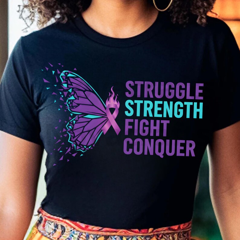 Puede incluir: Camiseta negra con un dise&ntilde;o de mariposa morada y turquesa y las palabras "STRUGGLE STRENGTH FIGHT CONQUER". La mariposa tiene un dise&ntilde;o de cinta y llama. La camiseta es de cuello redondo.