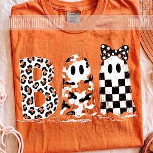 Peut inclure: Un t-shirt orange avec le mot BOO imprimé graphiquement. La lettre B présente un motif léopard, tandis que les deux O sont représentés par des fantômes. Un fantôme a un motif camouflage et l'autre un motif à damier noir et blanc.