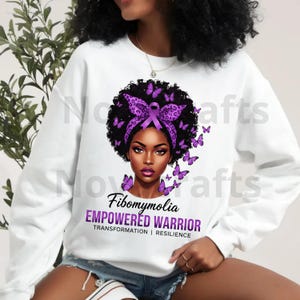 Puede incluir: Sudadera blanca con un gr&aacute;fico de una mujer adornada con una cinta morada y mariposas, con las palabras "Fibromialgia EMPOWERED WARRIOR". El dise&ntilde;o incluye las palabras "TRANSFORMATION | RESILIENCE".