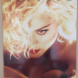 以下が含まれることがあります： 「MADONNA」の文字が金色で書かれたヴィンテージ雑誌の表紙。ブロンドの髪、赤いリップ、真剣な表情の女性のクローズアップです。全体的なカラースキームは暖かく、金、茶色、赤の色合いです。