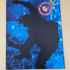 Peut inclure: Une affiche promotionnelle avec un fond bleu et une silhouette noire d'une personne en mouvement. Le texte "U2 ZOO TV OUTSIDE BROADCAST" est disposé en cercle, avec un dessin d'antenne parabolique.