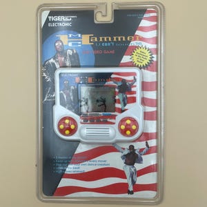 May include: A vintage Tiger Electronics MC Hammer U Can't Touch This video game, sealed in its original packaging. The game features a red, white, and blue design with images of MC Hammer. The packaging includes text and graphics.