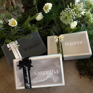 May include: Three gift boxes with names printed on them, surrounded by greenery and white flowers. The boxes are grey, white, and light grey, with ribbons and the names "William", "Reganne", and "Samantha" printed on them.