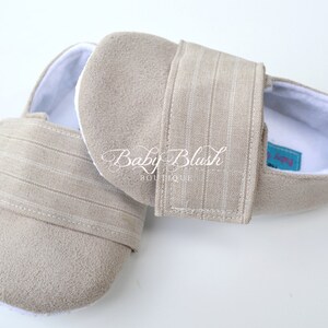 May include: A pair of beige baby shoes with a soft, suede-like texture. The shoes have a white interior and a striped strap closure. The text "Baby Blush Boutique" is visible on the side.