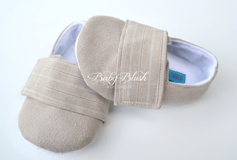 May include: A pair of beige baby shoes with a soft, suede-like texture. The shoes have a white interior and a striped strap closure. The text "Baby Blush Boutique" is visible on the side.
