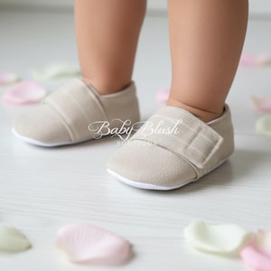 May include: Close-up of a baby's feet wearing beige fabric shoes with a velcro strap. The shoes have a white sole. Scattered around the feet are pink and white flower petals. The text "Baby Blush Boutique" is visible on the shoes.