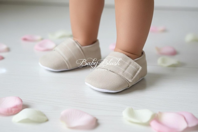 May include: Close-up of a baby's feet wearing beige fabric shoes with a velcro strap. The shoes have a white sole. Scattered around the feet are pink and white flower petals. The text "Baby Blush Boutique" is visible on the shoes.