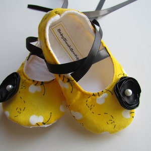 Puede incluir: Un par de zapatos de bebé amarillos con un patrón de abejas blancas y lazos de cinta negra. Los zapatos tienen un adorno de flor negra con un centro de perla. La etiqueta en el zapato dice "Baby Blush Boutique."