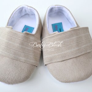 May include: A pair of beige baby shoes with a soft suede-like texture. Each shoe has a wide strap with horizontal stripes across the top. The interior is white, and a small label is visible inside each shoe. The words "Baby Blush Boutique" are written on the shoes.