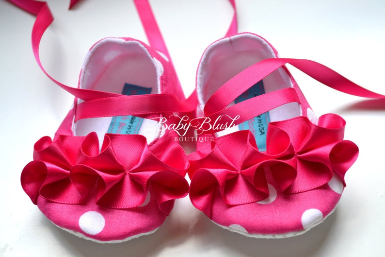 May include: Pink baby shoes with white polka dots and a pink satin bow. The shoes have a white lining and a pink ribbon tie.