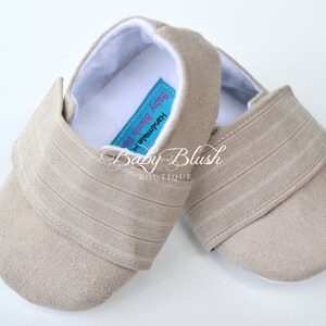 May include: A pair of beige baby shoes with a soft, suede-like texture. The shoes have a striped fabric detail on the strap and a white interior. The label reads "Baby Blush Boutique".