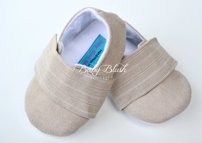 May include: A pair of beige baby shoes with a soft, suede-like texture. The shoes have a striped fabric detail on the strap and a white interior. The label reads "Baby Blush Boutique".