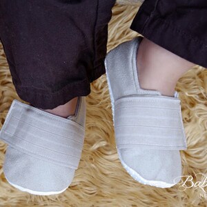 May include: A pair of light gray baby shoes with a hook and loop closure. The shoes are made of a soft material and have a white sole. The shoes are on a fluffy, light brown rug. The Baby Blush Boutique logo is visible.