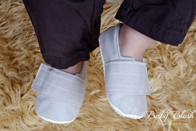 May include: A pair of light gray baby shoes with a hook and loop closure. The shoes are made of a soft material and have a white sole. The shoes are on a fluffy, light brown rug. The Baby Blush Boutique logo is visible.