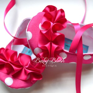 May include: Pink baby shoes with white polka dots and a large pink satin bow. The shoes have pink satin laces and a white lining. The shoes are labeled "Baby Blush Boutique".