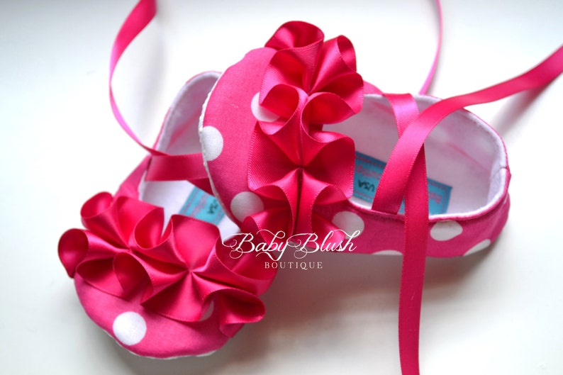May include: Pink baby shoes with white polka dots and a large pink satin bow. The shoes have pink satin laces and a white lining. The shoes are labeled "Baby Blush Boutique".