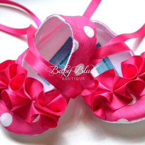 May include: Pink baby shoes with white polka dots and a large pink bow on the front. The shoes have pink satin ribbons that tie around the ankles.