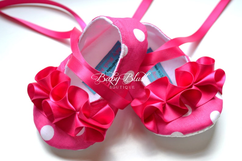 May include: Pink baby shoes with white polka dots and a large pink bow on the front. The shoes have pink satin ribbons that tie around the ankles.