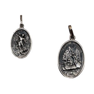 May include: Two silver-toned oval pendants. One pendant features an image of St. Michael, with the text "ST. MICHAEL". The other pendant has an image of an angel with the text "GUARD. ANGEL". Both pendants have a loop at the top for a chain.