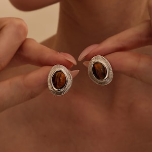 May include: A pair of stud earrings featuring oval-shaped tiger's eye gemstones set in silver-toned metal. Each earring has a textured, sunburst-style border surrounding the central brown and gold striped stone, creating a distinct metallic frame.
