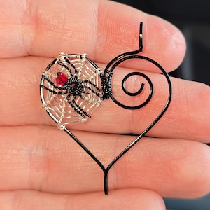 May include: A handcrafted pendant featuring a black wire heart with a spider and web design. The spider has a red bead body and is centered on a silver web. The pendant is a unique, gothic-inspired accessory.