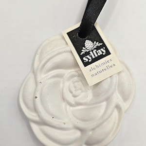 Könnte beinhalten: Ein weißer, rosenförmiger Lufterfrischer mit schwarzem Band. Das Etikett lautet "sylfay alchimies naturelles". Der Lufterfrischer ist ein dekorativer Artikel, wahrscheinlich für ein Haus oder Auto.