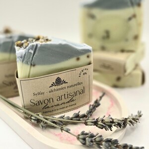 Puede incluir: Barras de jabón artesanal de lavanda con un diseño en capas, con una capa superior gris, un medio amarillo claro y una base blanca. El jabón está empaquetado con una etiqueta de papel marrón y ramitas de lavanda secas.